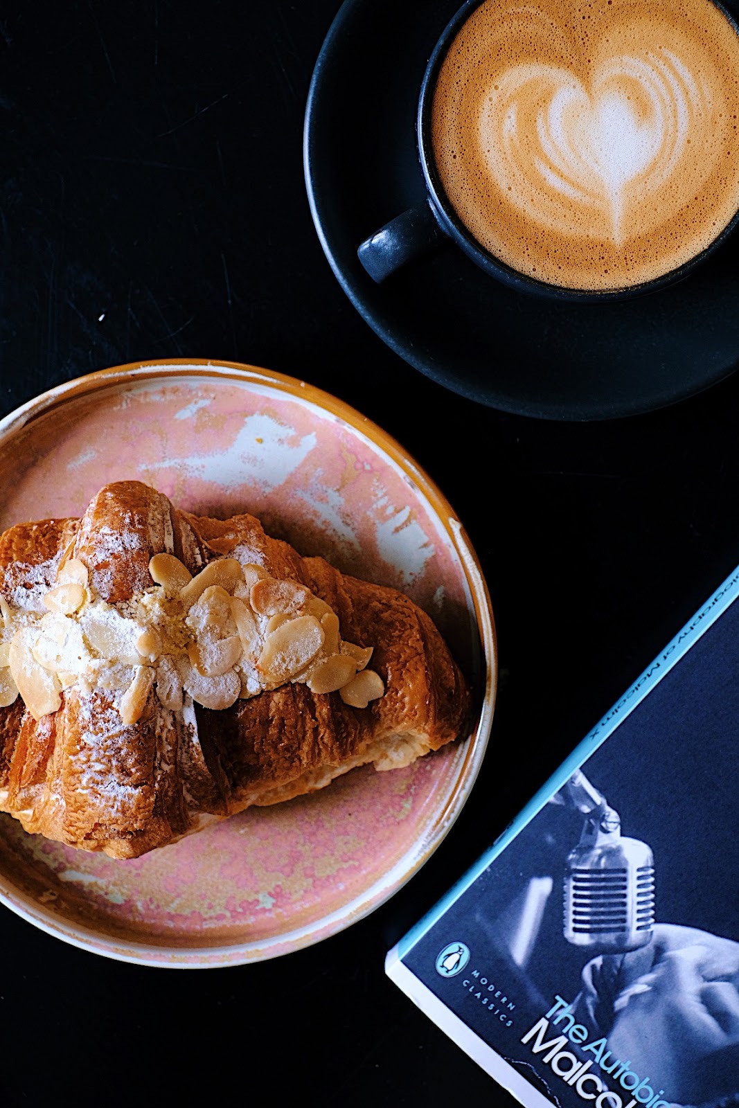 A latte with heart latte art beside an almond croissant on a pink plate